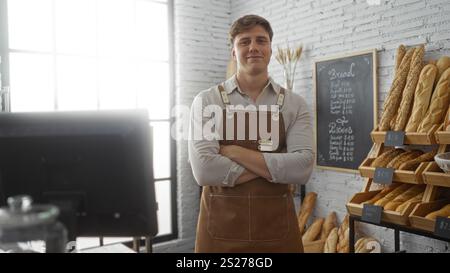Young man standing confidently in a bakery interior, posing with arms crossed in front of shelves stocked with various breads, creating a welcoming sh Stock Photo