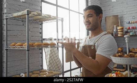 Young hispanic man in a bakery shop smiling while standing near a rack of freshly baked goods indoors Stock Photo