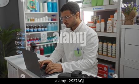 Young hispanic man working on a laptop inside a pharmacy, surrounded by shelves filled with medicine and supplies, wearing a white lab coat and glasse Stock Photo