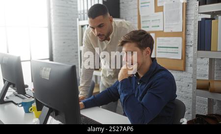 Two men collaborating at a computer in an office, one standing and one sitting, focusing on a work task, surrounded by documents and books in a profes Stock Photo