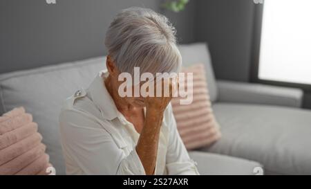 Grey-haired woman with short hair in an indoor living room setting, appearing pensive and covering her face, sitting on a couch, conveying emotions su Stock Photo
