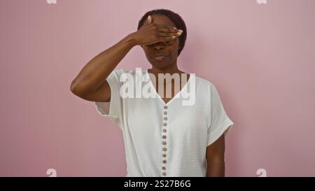 Young african american woman in a white shirt covering her eyes standing against an isolated pink background. Stock Photo