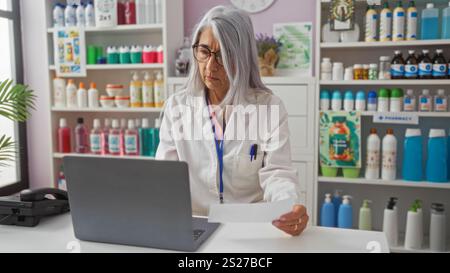 Grey-haired female pharmacist working on a laptop in a well-stocked pharmacy while holding a piece of paper, wearing a white lab coat with shelves of Stock Photo