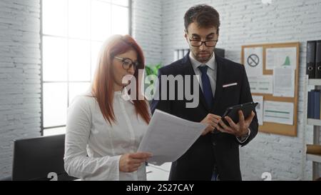 Business partners discussing documents in an office setting with man using tablet and woman holding papers, showcasing a collaborative work environmen Stock Photo