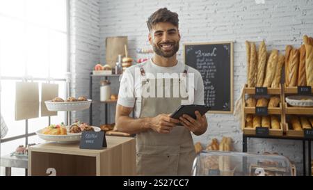 Young man with a beard working in a bakery shop, holding a tablet, surrounded by various types of bread and pastries displayed on shelves and counters Stock Photo