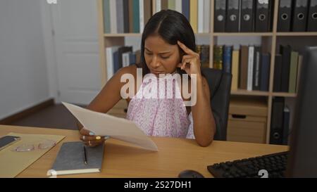 Woman concentrating on a document in an office setting, dressed in a patterned top, with a bookshelf in the background. Stock Photo