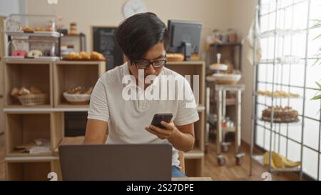 Handsome young chinese man using smartphone and laptop indoors in a bakery shop with pastries displayed on shelves. Stock Photo