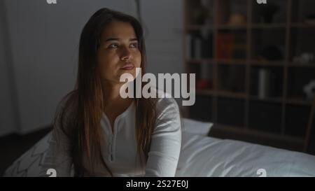 Woman sitting on bed in a bedroom with thoughtful expression, brunette hair, wearing white shirt, indoors with bedroom furniture, suggesting deep cont Stock Photo