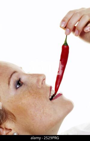 A young woman bites into a chilli pepper. Against a white background Austria Stock Photo