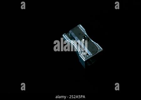 A close-up of a metal pencil sharpener against a dark, black background, showcasing its sleek and minimalist design. Ideal for projects related to education, stationery, office supplies, and creativity. The contrasting background emphasizes the sharpener's metallic texture, making it perfect for advertisements, academic materials, and artistic concepts Stock Photo