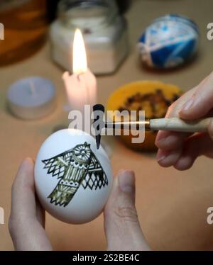 Process of painting an Easter egg. Bird stylized with an ornament on the egg. Photo. In hands is an egg, wax drawing tool. In background candles, wax, other eggs are out of focus. One candle burning. Stock Photo