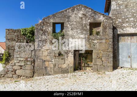 Front facade of an old abandoned granite house with pants growing out the window, front door is missing, blue sky. Stock Photo