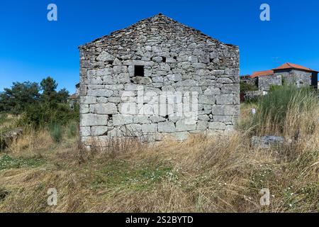 Side with window of an old abandoned granite detached house in Portugal with blue sky and tall grass. Stock Photo