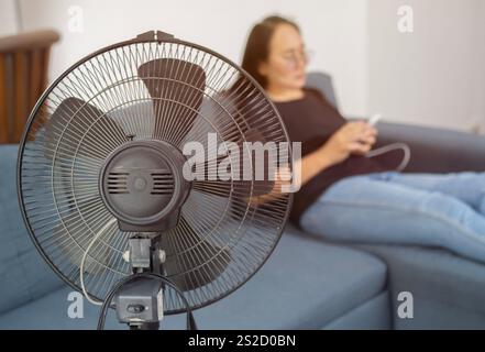 Close up shot of electric fan on floor at home. Closeup of black electric fan heater on floor in living room, with woman on couch in background. Clima Stock Photo