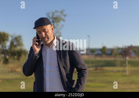 A middle-aged man strolls through a lush park, chatting on his mobile phone as he enjoys the bright daylight. Dressed casually Stock Photo