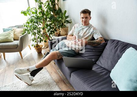 A young handsome man sits comfortably on a couch, enjoying coffee while working on a laptop. Stock Photo