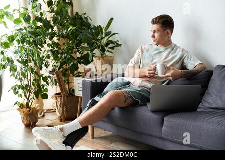 A handsome young man relaxes on a sofa, sipping coffee and using his laptop in a bright room. Stock Photo