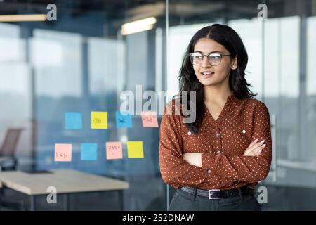 Young successful female office worker at workplace smiling with crossed arms looking towards the window. Businesswoman is satisfied working inside the office. Stock Photo