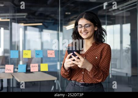 Young businesswoman successful office worker standing with phone in hands, typing message smiling happily. Woman in casual clothes standing by window. Stock Photo