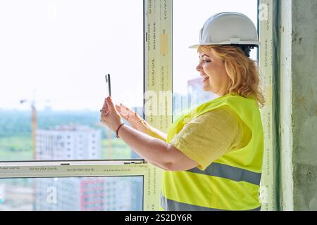 Female industrial worker in protective vest helmet using smartphone at construction site Stock Photo