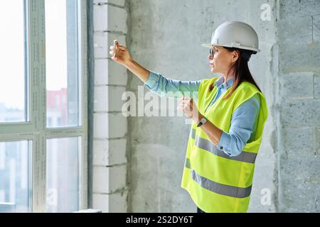 Female industrial worker in protective vest helmet using smartphone at construction site Stock Photo