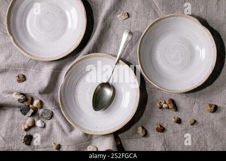 Set of empty craft ceramic white speckled bowls plates standing on grey linen table cloth with spoon and sea stones. Top view Stock Photo