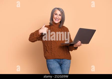 Smiling elderly woman in a brown sweater holding a laptop, gesturing a thumbs-up against a beige background Stock Photo