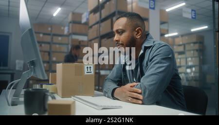 African American warehouse clerk scans parcel with barcode scanner, uses computer, works in post office. Sorting center worker carrying boxes for delivery to customers. Logistics distribution center. Stock Photo