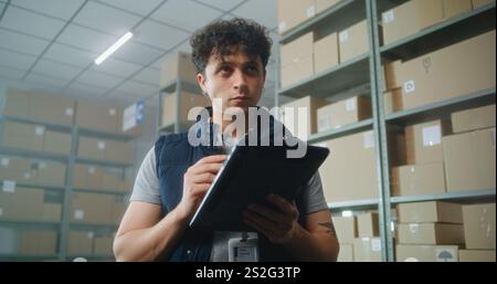 Male logistics specialist uses tablet computer and digital pencil, checks inventory. Warehouse multiethnic employees carrying cardboard boxes. Postal sorting center, E-commerce store delivery service. Stock Photo