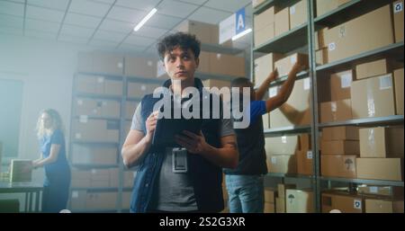 Male logistics specialist uses tablet computer and digital pencil, checks inventory. Warehouse multiethnic employees carrying cardboard boxes. Postal sorting center, E-commerce store delivery service. Stock Photo