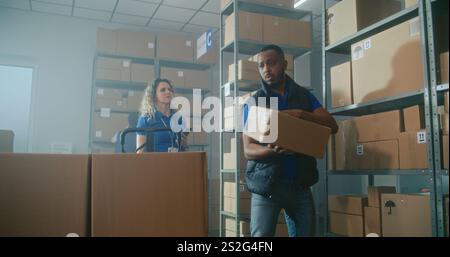 Female logistics specialist scans code on cardboard box using smartphone. African American warehouse worker loads parcel on pallet truck. Team of diverse warehouse employees working in sorting center. Stock Photo