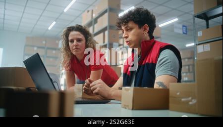 Two warehouse workers or logistics specialists scan parcel with scanner, prepare order for shipping to client, check information on laptop computer, work in post office. Online store delivery service. Stock Photo