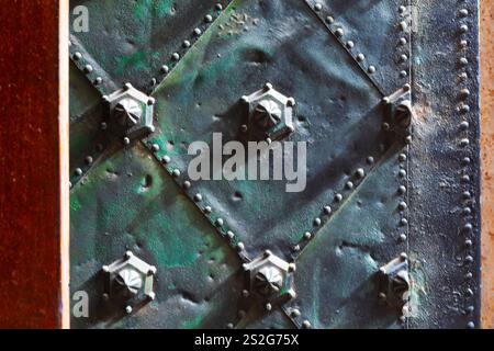 Weathered metal surface with a diamond pattern, featuring rivets and raised hexagonal bolts at intersections. Aged texture and patina of metal give it Stock Photo