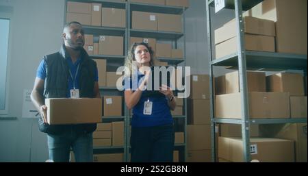 Female inventory manager walks with colleague, uses tablet computer. African American warehouse worker puts parcel on the shelf. Team of multiethnic employees working in logistics distribution center. Stock Photo