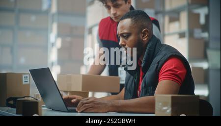 African American logistics specialist works on laptop computer in post office. Warehouse employee brings parcel to check delivery information. Sorting center of E-commerce online store. Slow motion. Stock Photo
