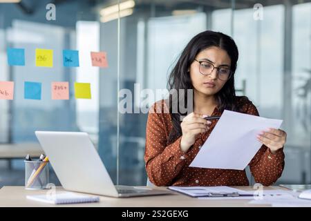 Serious focused woman behind paperwork reviewing analyzing and checking financial reports, financier accountant thinking and seriously concentrated at workplace inside office using laptop. Stock Photo