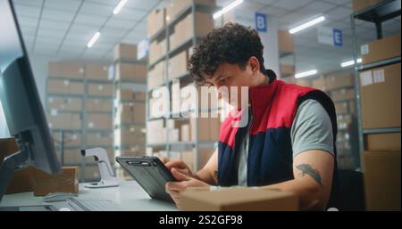 Male warehouse clerk scans parcels using tablet computer, works in postal service or online store logistics storage. Sorting center employees carrying cardboard boxes for delivery. E-commerce products Stock Photo