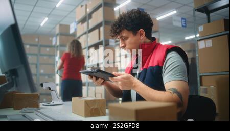 Male warehouse clerk scans parcels using tablet computer, works in postal service or online store logistics storage. Sorting center employees carrying cardboard boxes for delivery. E-commerce products Stock Photo