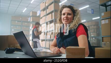 Portrait of female post office employee scanning cardboard parcel with barcode scanner, using laptop, looking at camera. Sorting center workers checking boxes for delivery. Warehouse of online store. Stock Photo