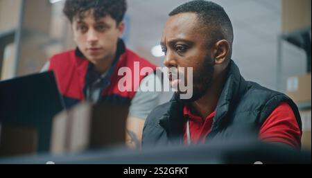 Close up of African American logistics specialist working on laptop in postal service. Male warehouse associate brings parcel to check delivery information. Sorting center of online store. Slow motion Stock Photo