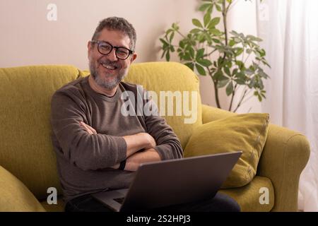A happy mature man wearing glasses and a casual outfit, sitting on a yellow sofa with a laptop, enjoying a relaxed moment in a living room. Stock Photo