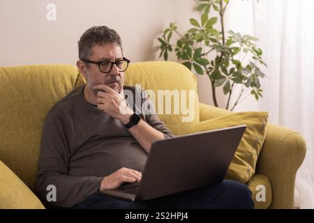 A mature man wearing glasses sitting on a yellow sofa, using a laptop with a thoughtful expression, in a cozy living room setting. Stock Photo