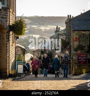 Tourists stroll up the steep Main Street in Haworth, with the scenic moors stretching into the distance on a sunny winter day. Stock Photo