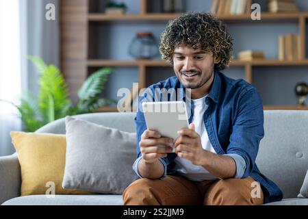 Young man sitting on sofa in living room at home with tablet computer in hands, smiling happily, using online app, browsing social media. Stock Photo