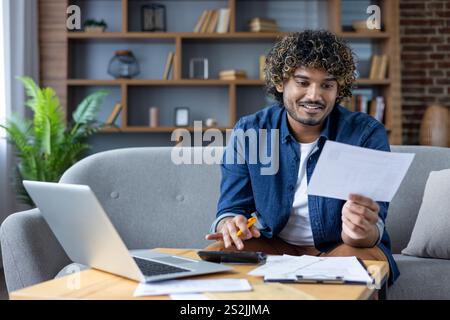 Young man doing paperwork sitting in living room of apartment on couch, paying credit checks and rent payments, using laptop and calculator. Stock Photo