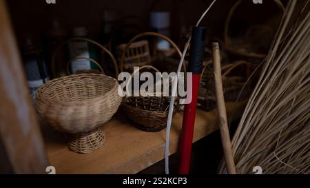 A collection of handcrafted wicker baskets displayed on a wooden shelf, accompanied by traditional tools and dried reeds for basket weaving. Stock Photo