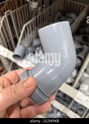 A person holds a grey plumbing elbow fitting in a hardware store aisle filled with various supplies. Bright lighting highlights the organization of th Stock Photo