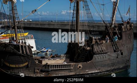 A replica of the ship “Santa Maria” docked in a sunny harbor, with tourists onboard. The wooden vessel showcases historic maritime design against Stock Photo