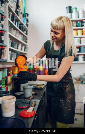 Smiling female professional graphic technician standing at printing shop and opening plastisol ink cans and choosing colors. Experienced print shop wo Stock Photo