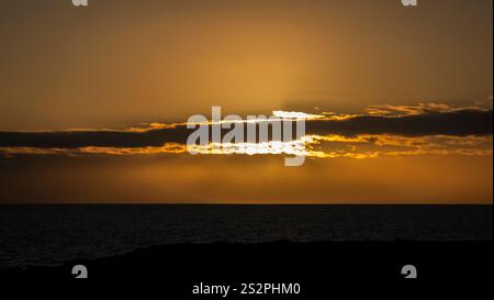Golden sunset over a calm ocean, with the sun partially hidden by dark clouds and silhouetted land in the foreground. Stock Photo